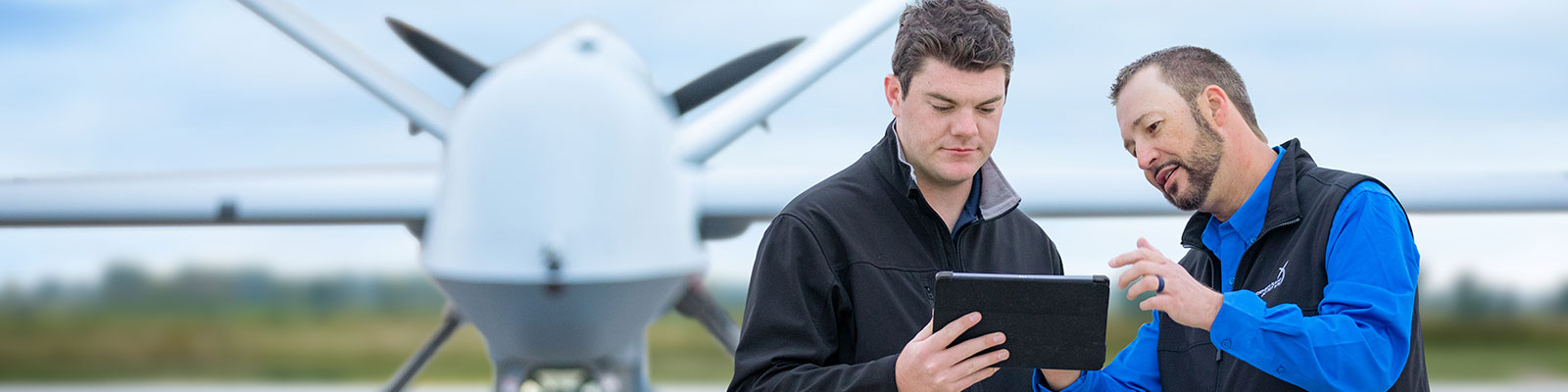 Two men look at a tablet device in front of a predator drone. 